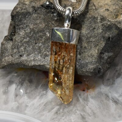 Amber crystal pendant with a silver cap hanging from a snake chain necklace on a dark rock surface, close-up detail side view.